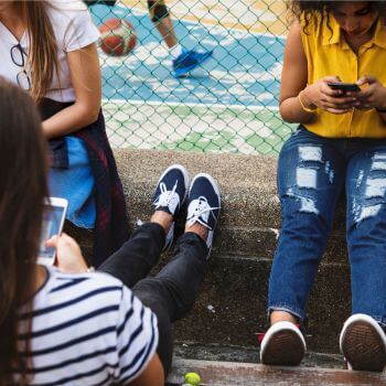 Three young people using smartphones and sitting on steps near a sports court.