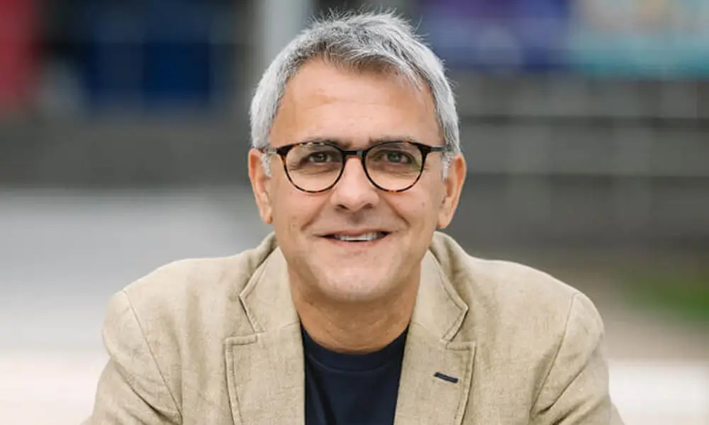 Portrait photo of Professor Daljit Nagra, a man in beige jacket with clasped hands sitting at a table outdoors.