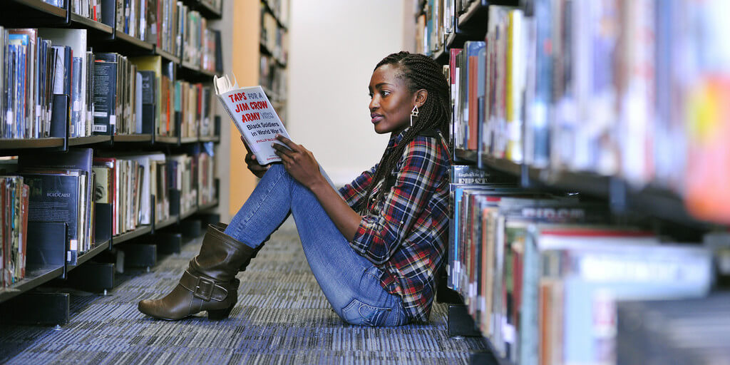 brunel student sitting on the floor in a library aisle reading a book