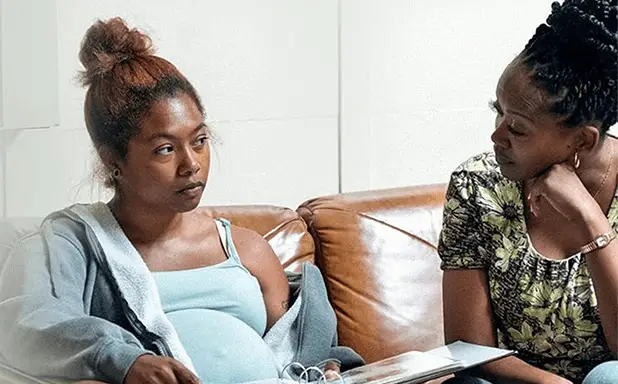 Two women sit closely on a brown couch, discussing something serious; one is visibly pregnant while holding a binder.