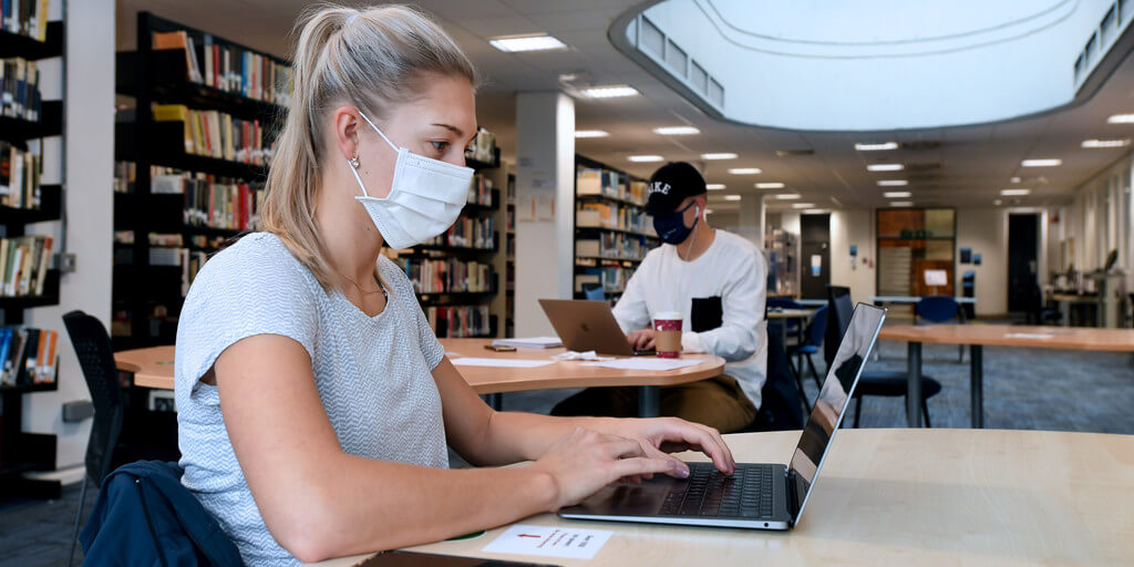 two students in face masks working on their laptops in the university library