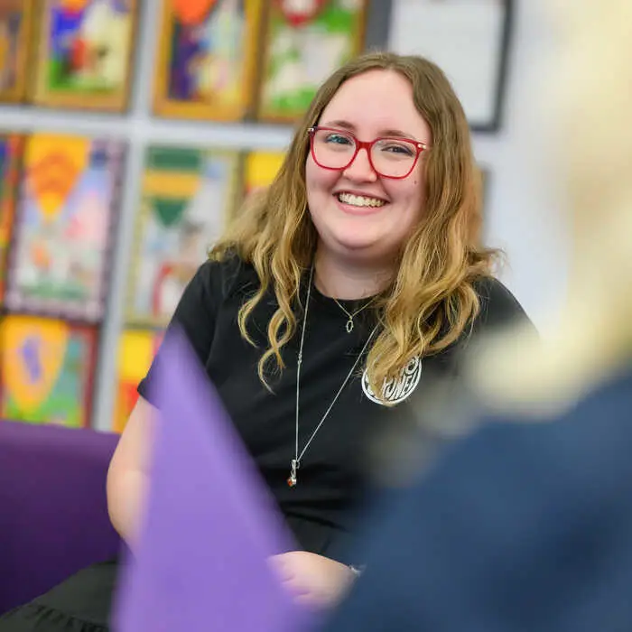 Smiling young woman with long hair and red glasses, sitting indoors with colorful artwork blurred in the background