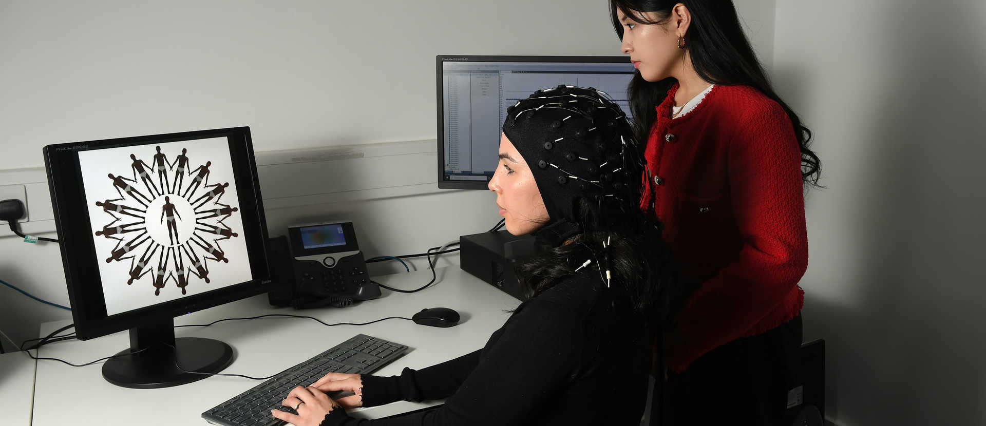 two female students sitting at a computer and one student is wearing brain wave scanning equipment