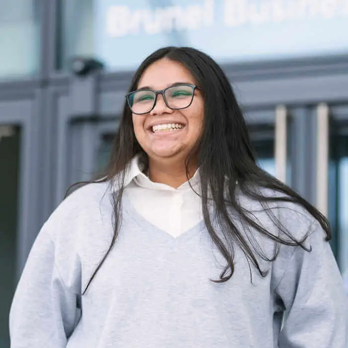 A person stands outside a building, wearing a light gray sweater and white shirt, against a backdrop of glass doors and a sign