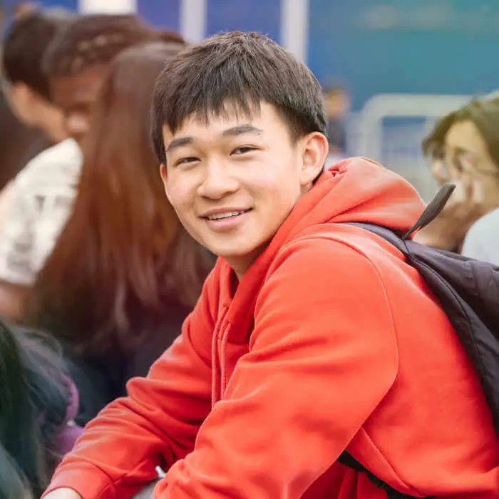 man in red jacket and with backpack sitting outdoors with people around
