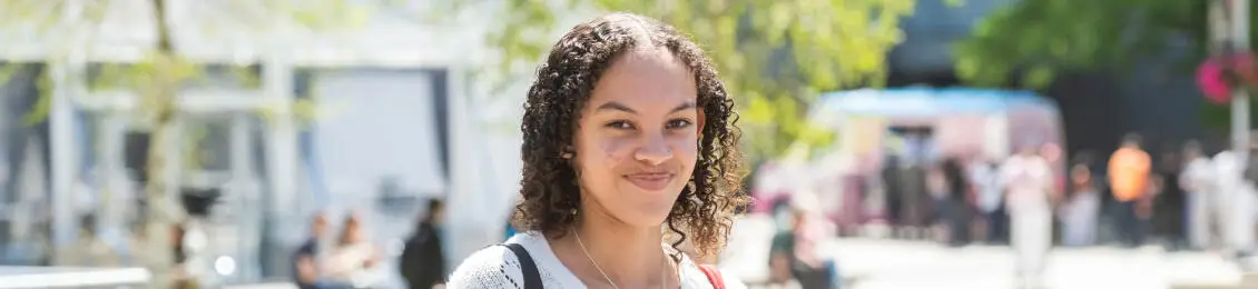 A woman with curly hair stands outdoors surrounded by a bustling crowd and greenery, enjoying a sunny day